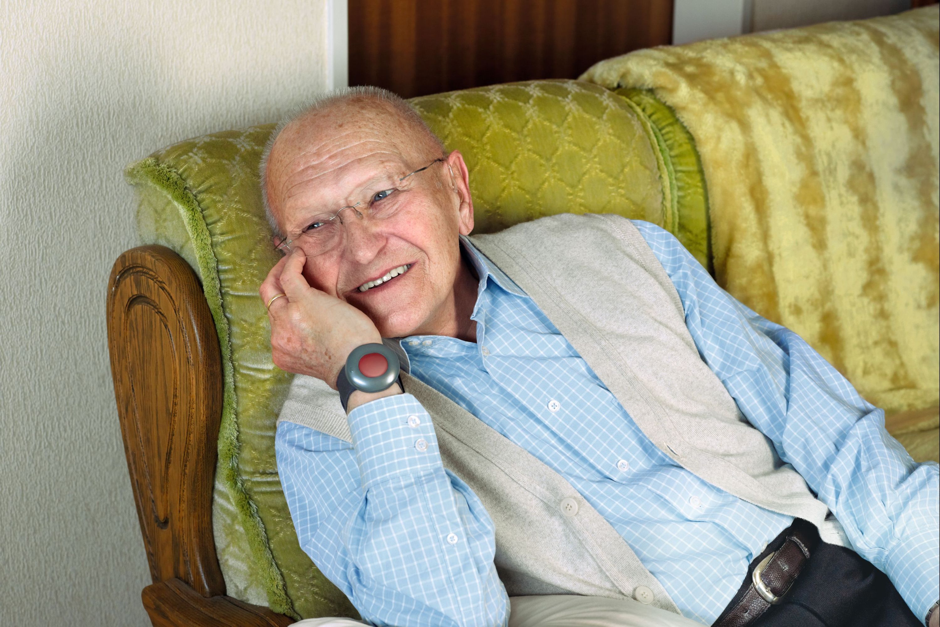 Ein älterer Herr sitzt auf seinem Sofa. Ums Handgelenk trägt er den Hausnotrufknopf des ASB.